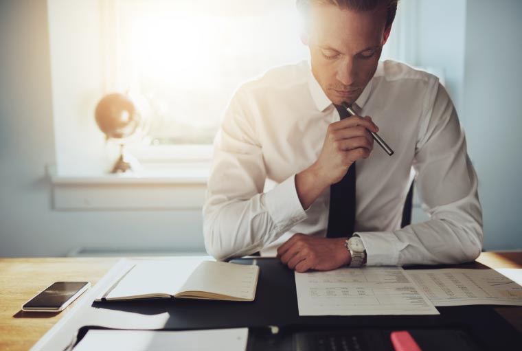 A focused businessman reviews documents at his desk, with sunlight streaming through the window beside him. He holds a pen to his chin in thought, surrounded by his phone, a notebook, and other office items. His associates are mentioned in the description of the scene.