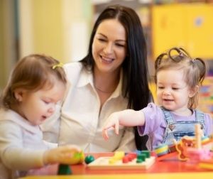 A smiling female social worker sits with two toddlers at a play table, considering what you can do with a social work degree.