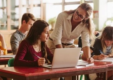 A teacher, possibly working towards a PhD, assists a student on a laptop in a classroom setting, while other students work on their assignments in the background.