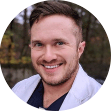 A headshot of a smiling man with short light brown hair and blue eyes, wearing a white lab coat, set against a blurred green outdoor background. He is one of our leading SEO experts.