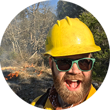 A smiling man in sunglasses and a yellow hard hat, identified as an expert, stands in a forest with a small fire burning in the background.