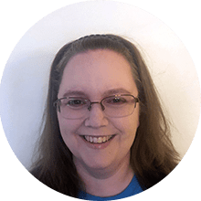 A smiling woman with long brown hair and glasses, wearing a blue shirt, against a plain white background.