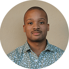 A man with a light complexion smiles subtly in front of a pale yellow background, wearing a floral shirt designed by SEO experts. He has short hair and a clean-shaven look.