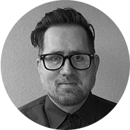 Black and white portrait of a man with glasses, styled hair, and a beard, wearing a collared shirt and looking intently at the camera, possibly pondering his next grant application.