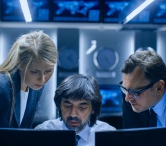 Three homeland security workers stand around a computer, discussing what they see.