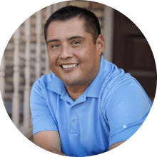 A smiling man wearing a blue polo shirt, sitting outdoors with a blurred background that suggests a casual environment. He appears content and relaxed, possibly reviewing ESL resources.