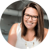 A cheerful woman with brown hair and glasses, wearing a white sleeveless top and a necklace, smiling at the camera in an indoor, casually professional college setting.