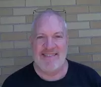 A smiling bald man with glasses on his head, wearing a black t-shirt, standing in front of a brick wall.