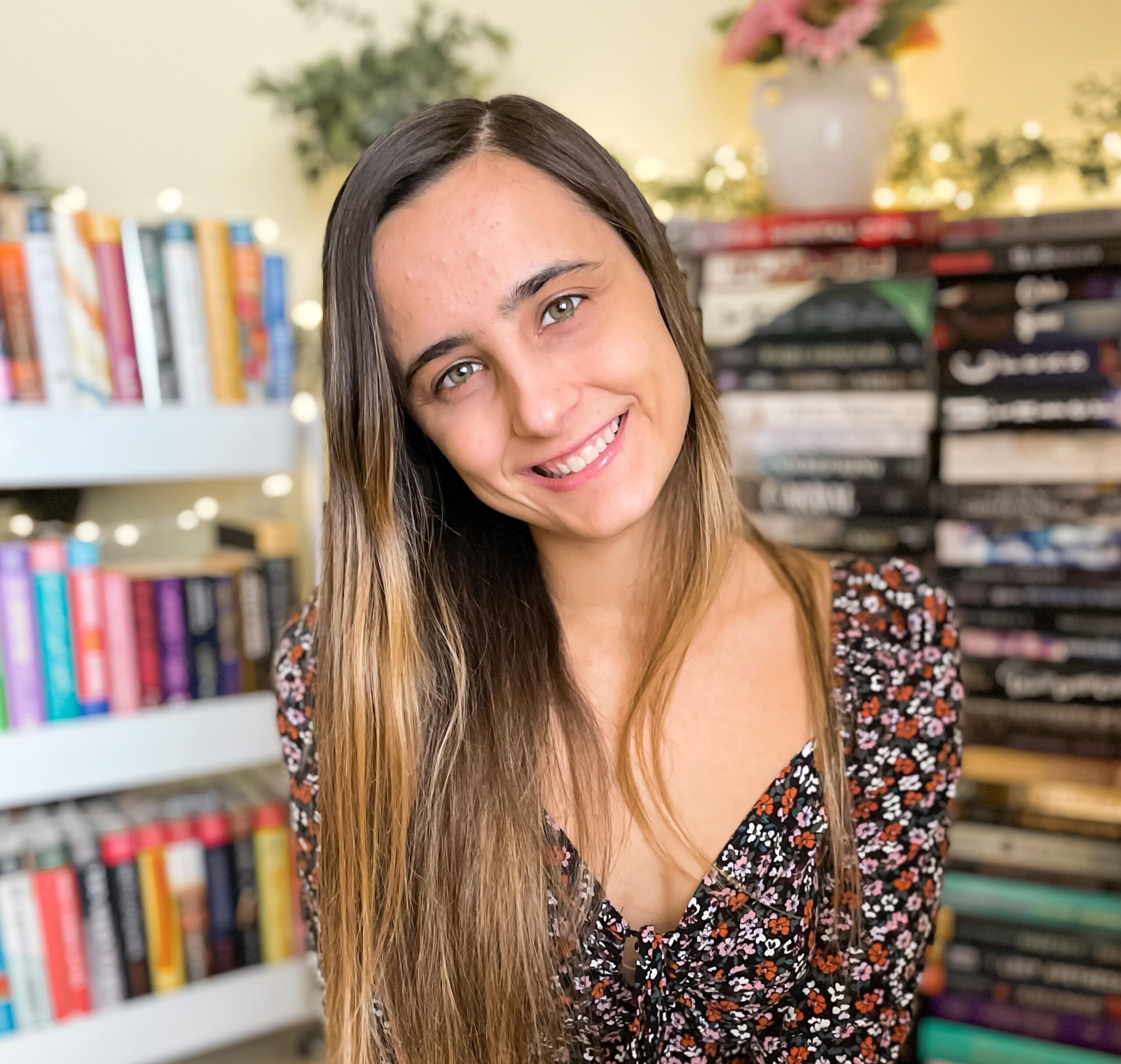 A smiling young woman with long, straight brown hair wearing a floral dress, sitting in front of a bookshelf filled with colorful books, with string lights and a vase of flowers in the background.