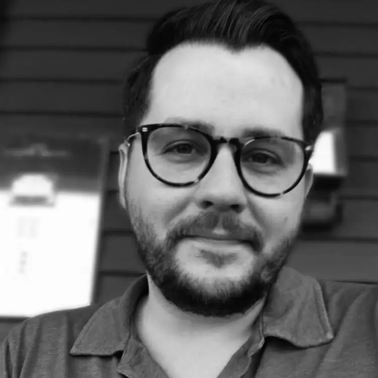 A black and white headshot of a man with glasses, sporting a beard and wearing a collared shirt, smiling slightly at the camera.