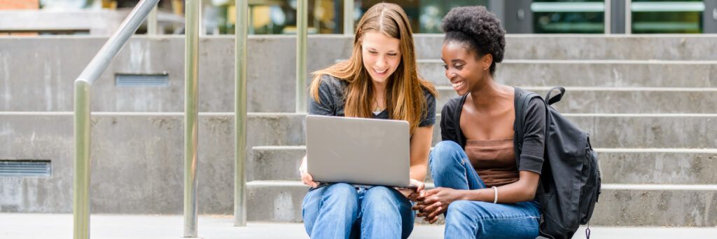 Two female students sitting on some stairs looking at a laptop.