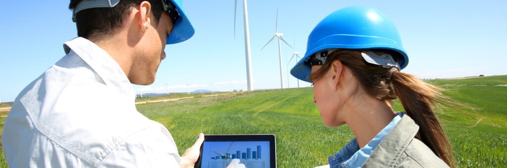 A male worker and female worker surrounded by wind turbines and looking at a computer.