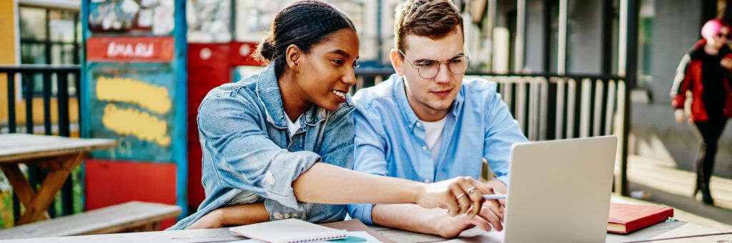 A male student and a female student sitting on a bench looking at work on a laptop.