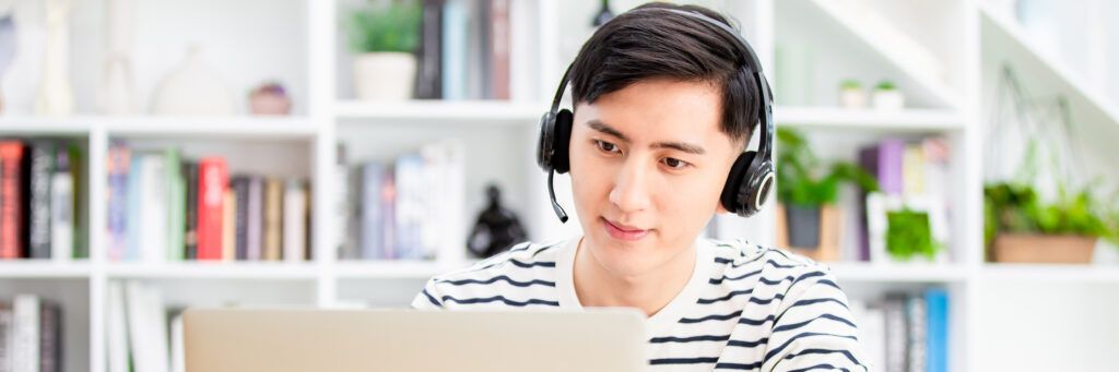 A male student in the library working on a laptop while wearing headphones.