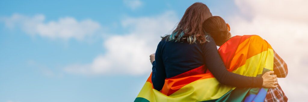 Two women wrapped up in a LGBTQIA flag looking at the sky.