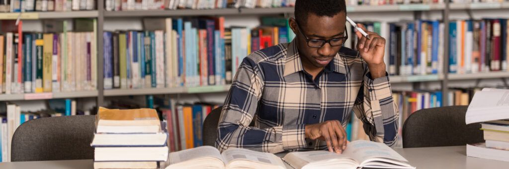 A Black male student studying from books in a library.