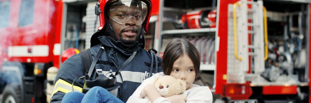 A fireman holding a little girl and her teddy bear in front of a fire truck.