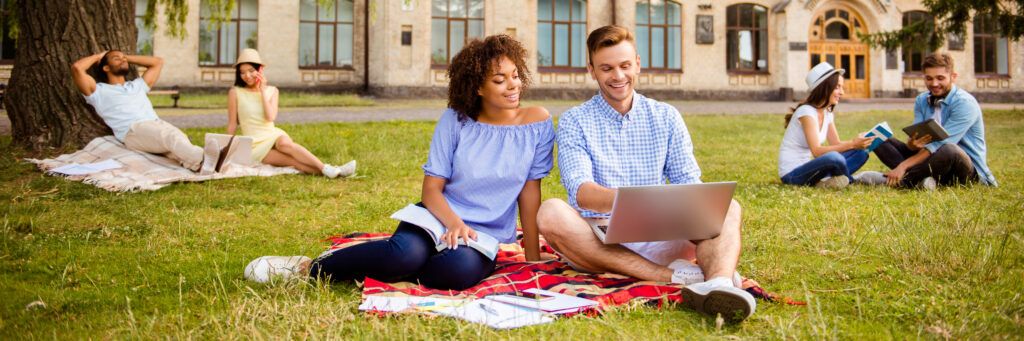 Two online students looking at a laptop while sitting on a blanket in the park.