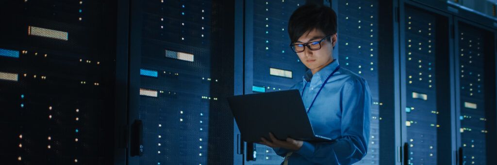 Worker in blue shirt views laptop while standing in front of bank of electronics.