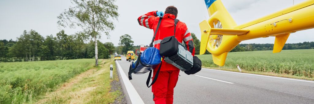 An emergency responder carries two duffel bags with equipment after landing in a helicopter on a remote road.