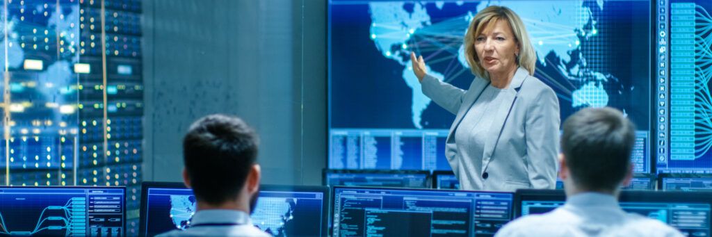 A woman points to a large screen with a map of the world as she lectures two young men sitting in a room full of computers.