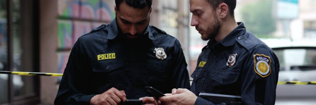 Two law enforcement offers are seen looking at their phones while standing in front of yellow police tape at a crime scene.