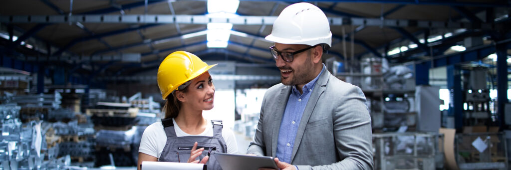 A young woman in a yellow hard hat and overalls talks to a supervisor while working in a warehouse.