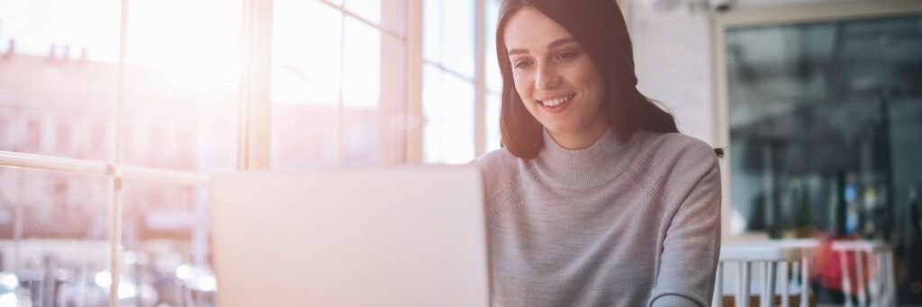 A young woman smiles as she looks at her computer screen while sitting in a public place.