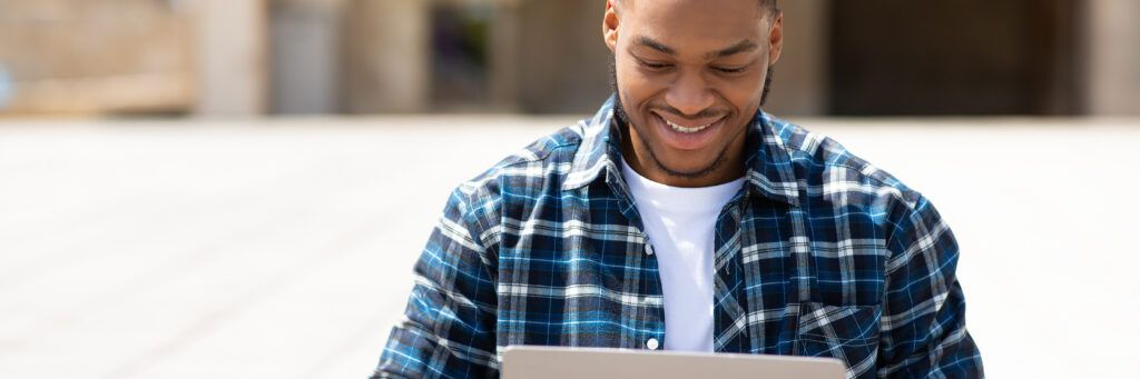 A young man in a blue flannel shirt smiles as he reads more about online colleges with open admission policies while sitting outside a college campus.