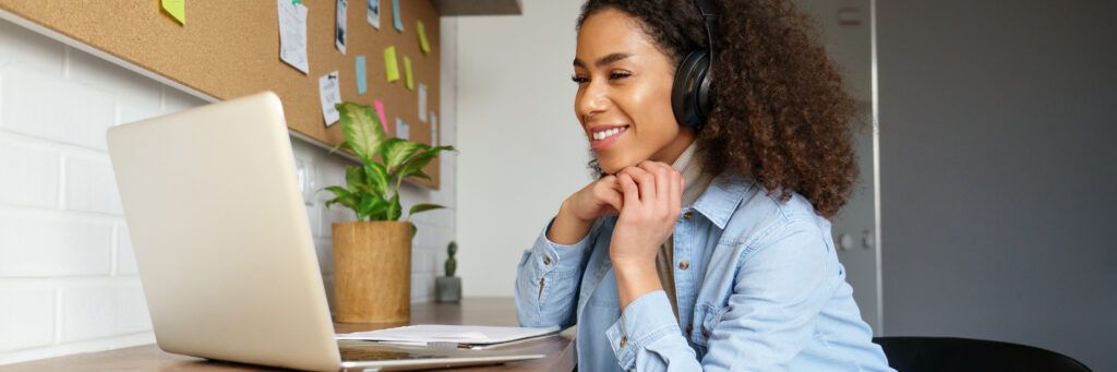 A young woman wearing headphones smiles as she researches online community colleges on her portable computer.