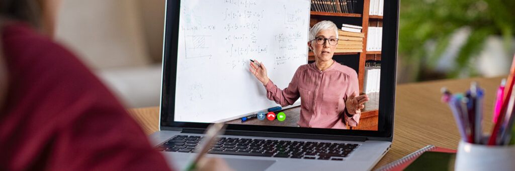 A student takes notes while watching a college professor explain a lesson plan on a whiteboard through a live video lecture on a laptop.