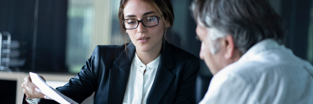 A woman in office attire holds out a document to show it to an older man sitting next to her.