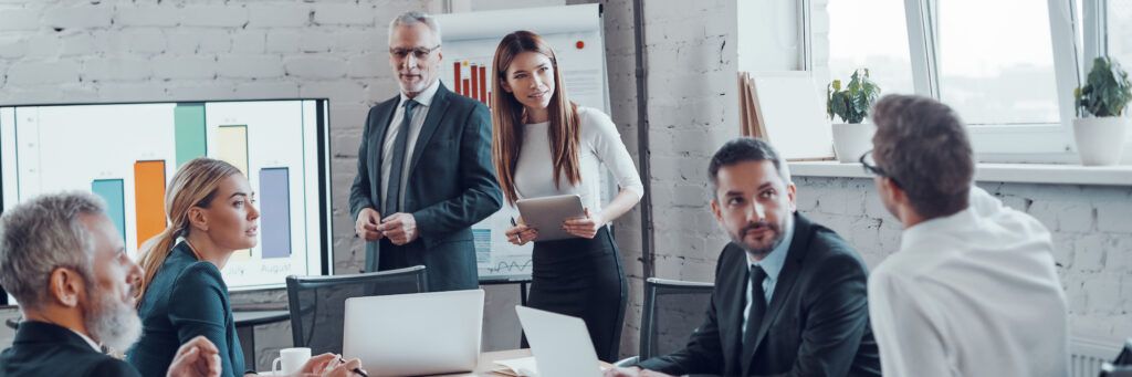 In an office, a group of men and women examine a graph on a large screen and talk about its implications.