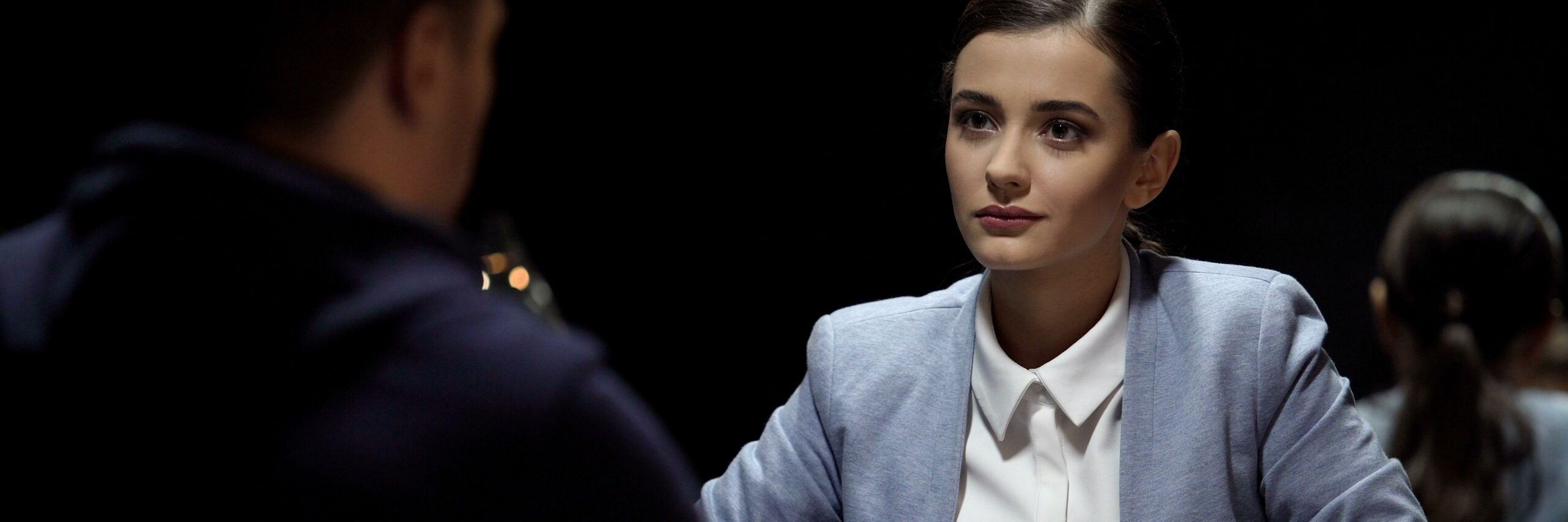 A young woman with a composed expression sits across from a man, engaging in a serious conversation in a dimly lit room. Another person sits in the background, partially visible.