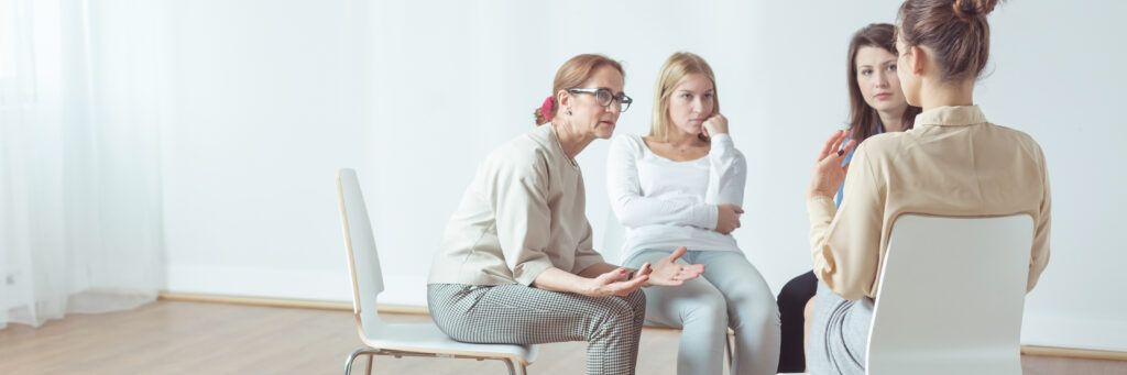 A group of woman are seen sitting in chairs in a circle while talking to each other in an empty-looking room.