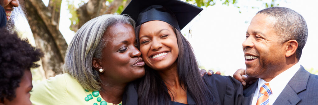 A young woman wearing a graduation cap and gown smiles and celebrates with her family after graduating from a top HBCU for online school. 