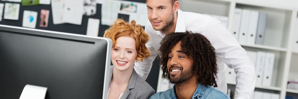 Two men and a woman smile and look at a computer together, choosing between an MPA vs MBA.