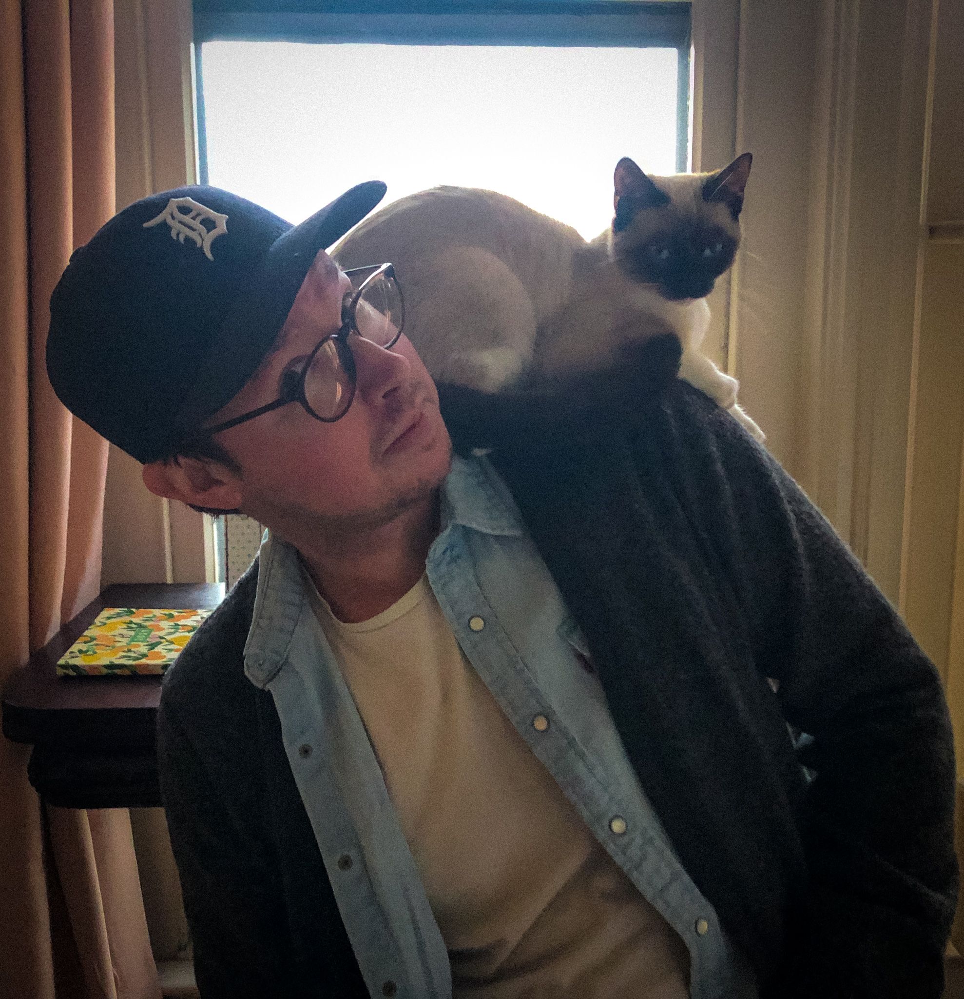 A man in glasses and a baseball cap looks to the side while a Siamese cat sits comfortably on his shoulder, both inside a softly lit room.