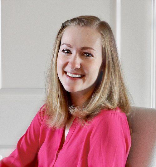 A smiling woman with blonde hair dressed in a bright pink blouse, sitting indoors with a soft-focus background.