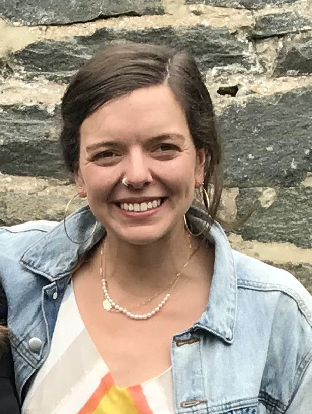 A smiling woman with brown hair wearing a denim jacket and pearl necklace standing in front of a stone wall.