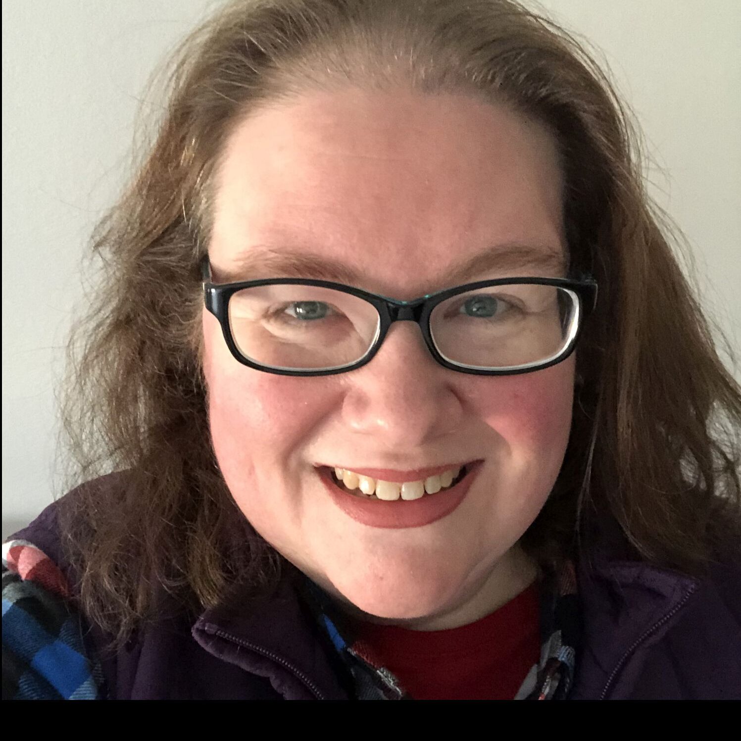 A smiling woman with long brown hair, wearing glasses and a purple jacket over a plaid shirt. She is in a room with a white wall.