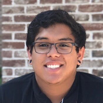 A close-up portrait of a smiling young man with glasses and short black hair, standing in front of a brick wall. He is wearing a black shirt.