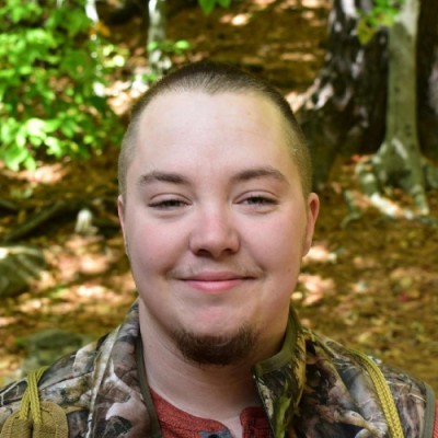 A smiling young man with a buzz cut, wearing a camouflage jacket, standing outdoors with trees and greenery in the background.