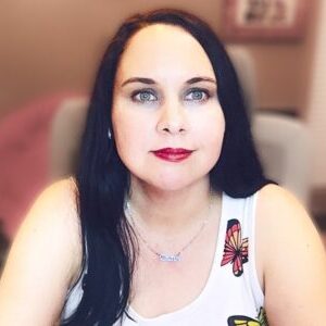 A woman with long black hair and fair skin wearing a white tank top with a butterfly design, sitting indoors with a softly blurred background. She looks directly at the camera, wearing red lipstick.
