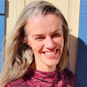 A smiling woman with long blonde hair, wearing a red and black top, standing against a sunlit blue wall.