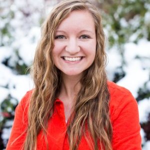 A young woman with long, wavy hair and a vibrant red shirt smiles warmly against a snowy background.