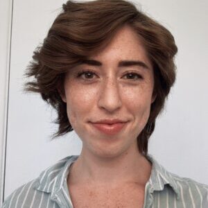 A portrait of a smiling woman with short, wavy brown hair and a light blue striped shirt. She has fair skin and is standing against a plain white background.