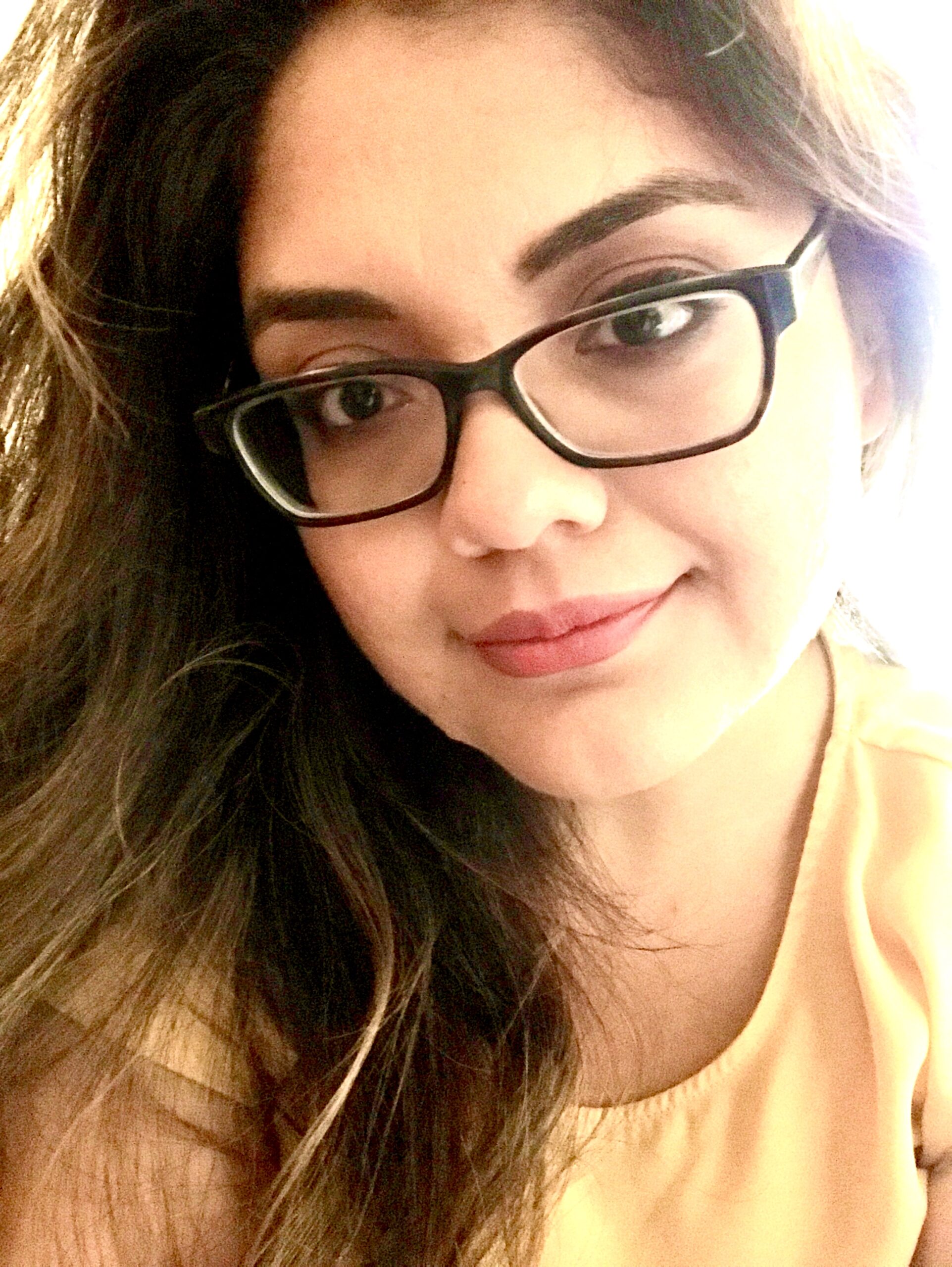 A close-up portrait of a woman with long, wavy dark hair wearing black glasses and a yellow blouse, smiling slightly at the camera.