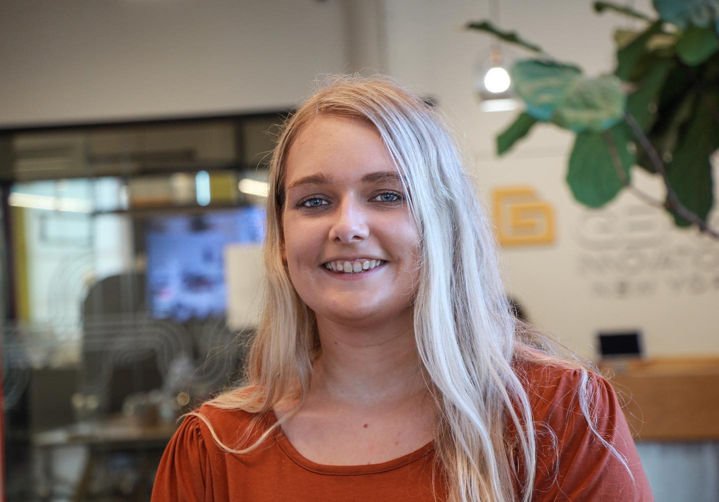 A young woman with long blonde hair and a bright smile, wearing an orange top, standing in an office environment with a blurred background featuring plants and a logo.