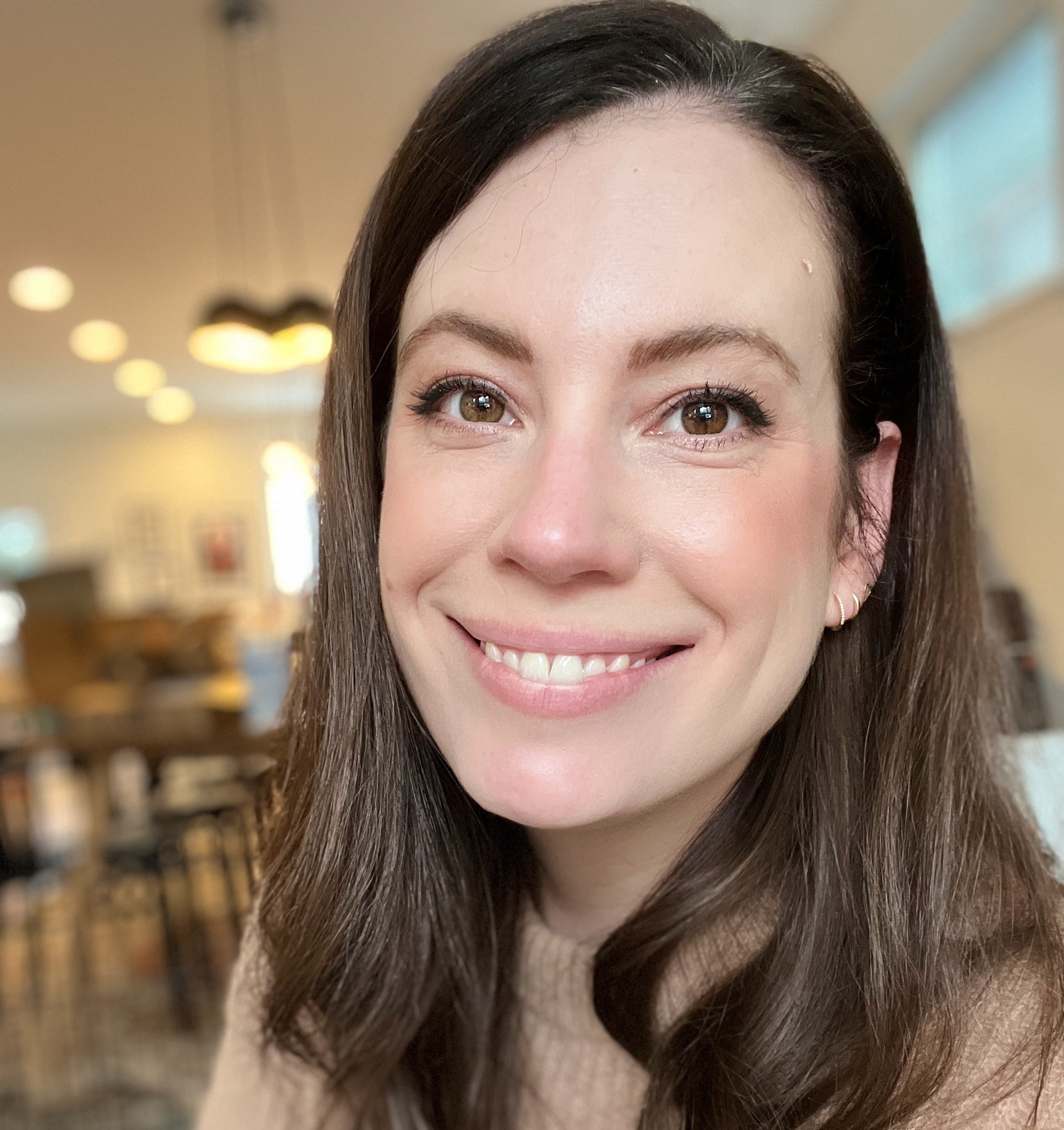 A close-up portrait of a smiling woman with long dark hair, wearing a beige sweater. She is in a warmly lit room with blurred chairs and hanging lights in the background.